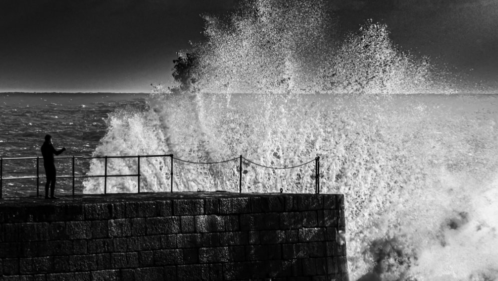 A shocked person standing on a stone pier as a large wave crashes against it.